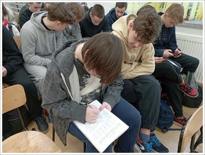 uczniowie siedzą na krzesełkach i piszą  na kartkach papieru