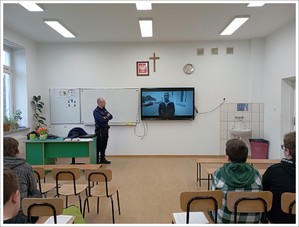 policjant stoi obok projektora, na ścianie wyświetlony jest obraz przedstawiający młodego człowieka w klasie na sali na krzesełkach siedzą uczniowie