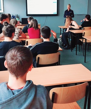 uczniowie siedzą w klasie w ławkach. przed nimi stoi policjantka. w tle biała tablica