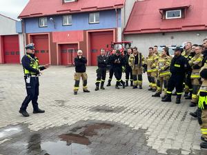 na placu przed budynkiem straży pożarnej stoją strażacy przed nimi stoi policjant ruchu drogowego