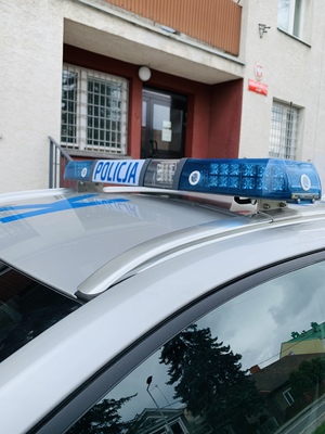 dach radiowozu policyjnego z policyjną belką i napisem policja w tle wejście do budynku