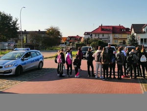 policjant stoi przed grupą młodzieży w tle budynek policyjny radiowóz oraz pojazdy