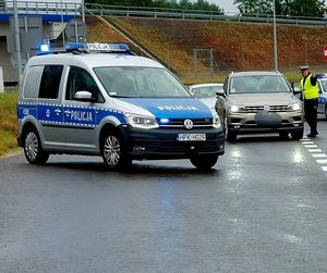 policyjny radiowóz obok radiowozu osobowy samochód obok którego stoi policjant