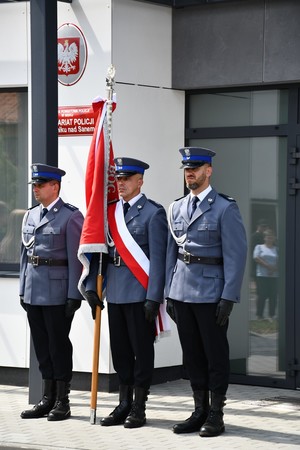 trzech policjantów ze sztandarem stoi przy budynku komisariatu