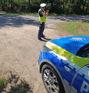 policjant stoi przy drodze prowadzi pomiar prędkości pojazdów w tle radiowóz