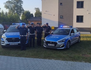 policjanci oddają honory stojąc obok dwóch radiowozów w których włączone są sygnały świetlne i dźwiękowe w tle budynek posterunku policji