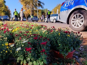 policjanci kierują ruchem na drodze w tle radiowóz oraz kwiaty na parkingu naprzeciw cmentarza