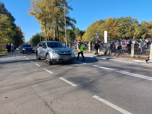 policjanci stoją na jezdni po której jada pojazdy oraz chodzą przechodnie z kwiatami