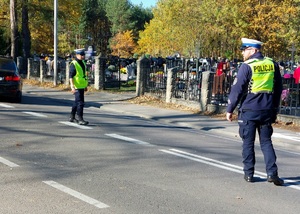 policjanci kierują ruchem na jezdni po której chodzą piesi i jadą pojazdy