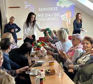 spotkanie z seniorami seniorzy otrzymują smycze od kobiety w tle stoi policjantka na ścianie napis seniorze nie daj się oszukać
