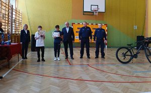 na sali sportowej stoi chłopiec z dyplomem obok niego kobieta, mężczyzna oraz dwaj policjanci przed nimi stoi rower