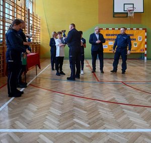policjant wręcza chłopcu puchar obok stoi kobieta, mężczyźni oraz policjanci przed nimi na hali sportowej stoi rower