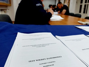 na stole leży kartka z napisem test wiedzy zawodowej eliminacje w tle policjanci rozwiązują test przy stole