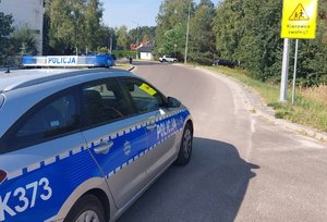policyjny radiowóz zaparkowany na poboczu drogi w tle budynek oraz znak drogowy ostrzegawczy z napisem kierowco zwolnij