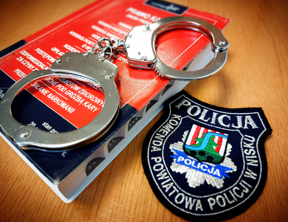 kajdanki leżą na kodeksie karnym który leży na stole. Obok leży emblemat z wyhaftowanym logo komendy powiatowej policji w nisku z napisem komenda powioatwa policji w nisku