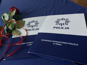 na ścianie napis komenda powiatowa policji w nisku na stole leżą teczki z aktami mianowania oraz róża