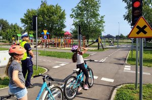 policjant ruchu drogowego stoi na boisku w otoczeniu dzieci z rowerami i w kaskach w tle budynek szkoły egzamin praktyczny  na karte rowerową