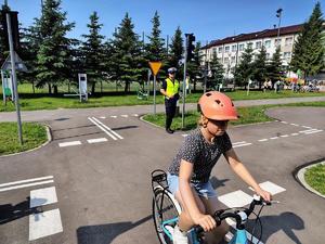 policjant ruchu drogowego stoi na boisku w otoczeniu dzieci z rowerami i w kaskach w tle budynek szkoły egzamin praktyczny  na karte rowerową