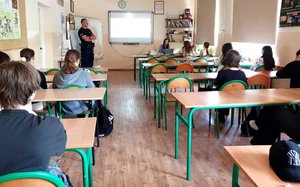 UCZNIOWIE SIEDZĄ W KLASIE W ŁAWKACH W TLE KOBIETA SIEDZI ZA BIURKIEM OMAWIA PREZENTACJĘ MULTIMEDIALNĄ PO DRUGIEJ STRONIE STOI POLICJANT