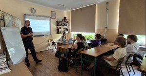 młodziez siedzi w szkolnych ławkach w klasie przy biurku kobieta obok stoi policjant prowadzi prelekcje
