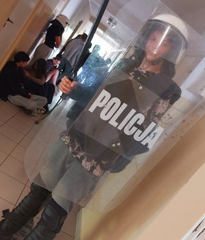 uczeń w korytarzu ubrany w zestaw przeciwuderzeniowy trzyma  tarczę z napisem policja na głowie ma kask w ręku pałkę szturmową