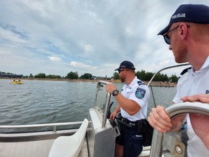 policjanci na łodzi motorowej na wodzie patroluja teren zbiornika wodnego na brzegu którego wypoczywają osoby
