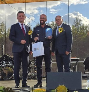 policjant stoi na scenie w rękach trzyma statuetkę obok stoi burmistrz oraz starosta