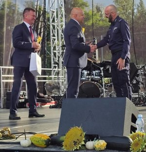 na scenie stoi policjant dwóch mężczyzn starosta oraz burmistrz podają mu dłonie w geście gratulacji