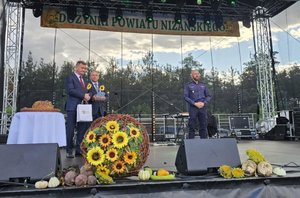 policjant oraz dwóch mężczyzn stoją na scenie w tle napis dożynki powiatu niżańskiego na scenie poukładane są słoneczniki oraz plony