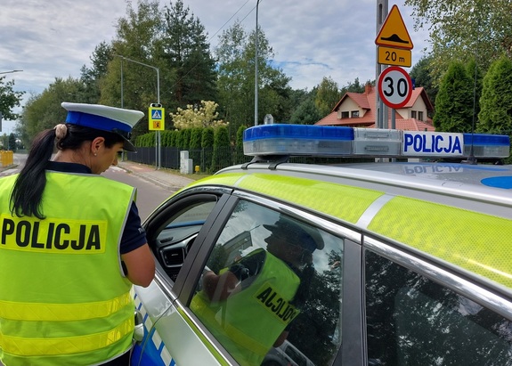 policjantka stoi przy radiowozie w tle jezdnia oraz budynek szkoły i znaki drogowe ograniczające prędkość