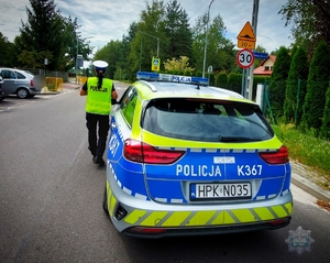 policjantka stoi przy radiowozie na drodze w tle znaki drogowe budynki i zaparkowane na poboczu pojazdy osobowe