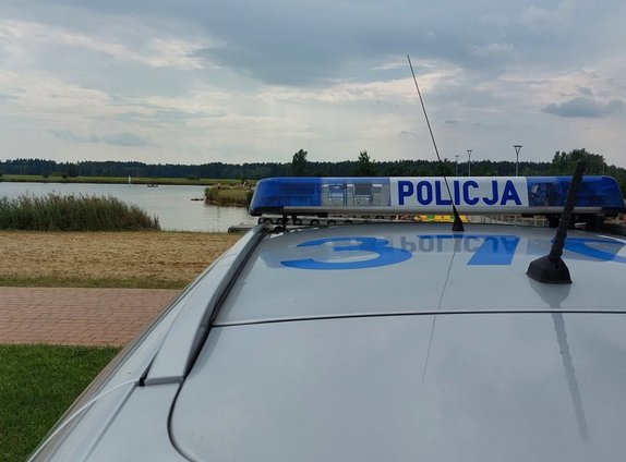 widok dachu radiowozu policyjnego z policyjną belką z napisem policja w tle widok zbiornika wodnego