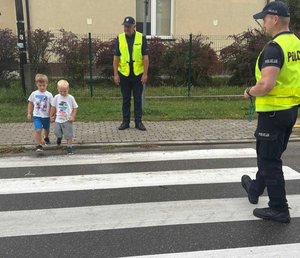 dzieci przechodzą przez jezdnie na przejściu dla pieszych  w tle policjanci
