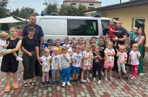 policjanci stoją z grupą dzieci przy radiowozie jeden z policjantów trzyma dziecko na rękach