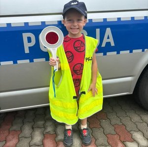 chłopczyk stoi przy radiowozie z tarczą do zatrzymywania pojazdów w kamizelce odblaskowej i policyjnej czapce