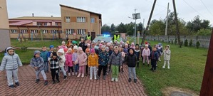 Grupa dzieci stoi wraz z policjantem na tle radiowozu.