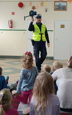 Policjant stoi przed dziećmi i trzyma w ręce urządzenie do pomiaru prędkości.