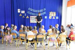 Policjantka stoi naprzeciwko siedzących na krzesełkach dzieci i mówi.