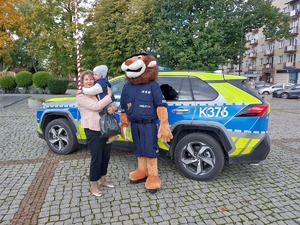 Maskotka policji stoi przy kobiecie trzymającej dziecko na rękach.
