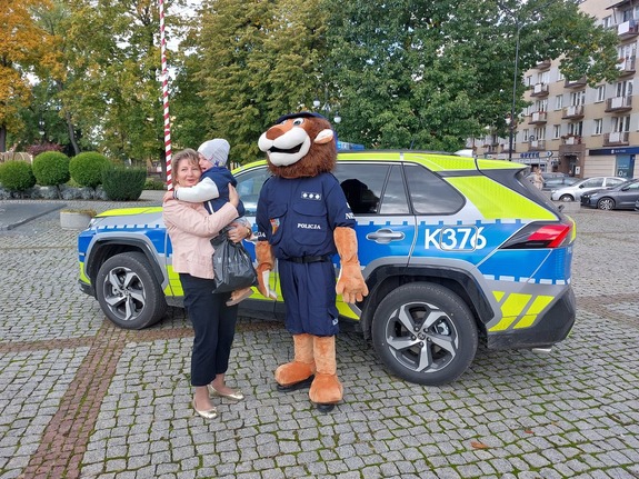 Maskotka policji stoi przy kobiecie trzymającej dziecko na rękach.