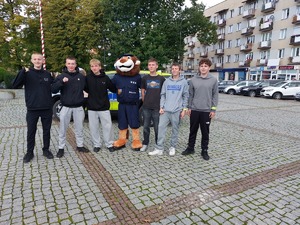 Młodzież stoi z maskotką policji na tle radiowozu i pomnika.