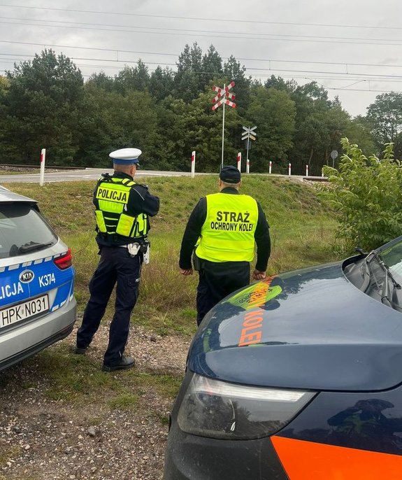 Policjant wraz z funkcjonariuszem Służby Ochrony Kolei stoją obróceni tyłem i patrzą na niestrzeżony przejazd kolejowy przy którym stoi Krzyż św. Andrzeja.