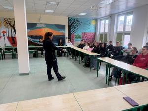 Policjantka stoi naprzeciwko uczniów, którzy siedzą na krzesełkach.