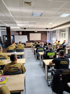 Strażacy siedzą w ławkach, policjant przy biurku prowadzi wykład