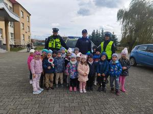 Grupa przedszkolaków stoi przed policjantami na tle radiowozu i szkoły.
