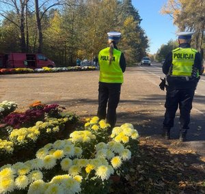 Policjanci stoja przy cmentarzu i nadzorują ruch. Na zdjęciu widać stoisko z chryzantemami