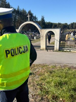 Policjant nadzoruje ruch w rejonie cmentarza. W tle widac cmentarną bramę