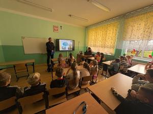 dzieci siedzą na krzesełkach, policjant stoi i omawia slajd wyświetlony na tablicy