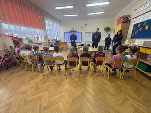 Dzieci siedzą na krzesełkach, policjanci stoją naprzeciwko i opowiadają