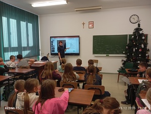 Policjantka stoi  i mówi, dzieci siedzą na krzesełkach i słuchają